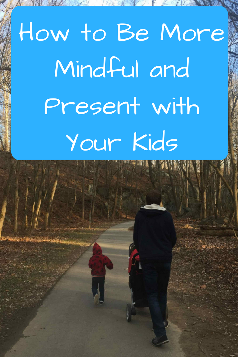 How to Be More Mindful and Present with Your Kids. (Photo: Man in a blue hoodie walking down a paved trail surrounded by trees pushing a stroller, with a child walking next to him.)