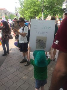 Photo from the back of a white child holding up a sign that says "I Love Trains" with other people on a sidewalk. 