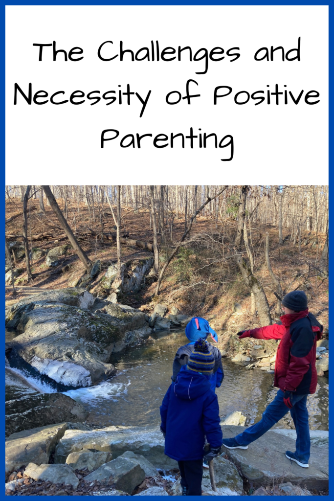 The Challenges and Necessity of Positive Parenting; photo of a man in a red winter coat next to a kid with a blue winter coat and hat standing next to a stream with a small waterfall with bare trees in the background