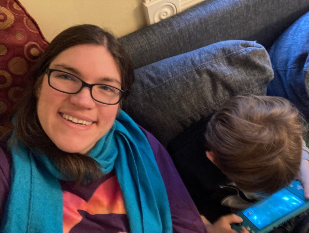 Shannon (a white woman in a teal scarf) looking up at the camera with my younger son (a white boy) sitting next to me playing a Nintendo Switch on a blue couch