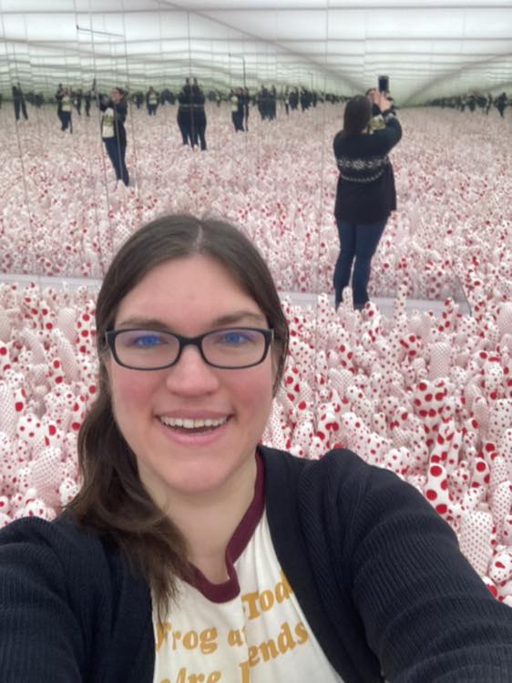 Selfie of Shannon (a white woman in a Frog and Toad shirt) in the Kusama exhibit of white stuffed ovals with red polka-dots and mirrors upon mirrors reflecting all of it