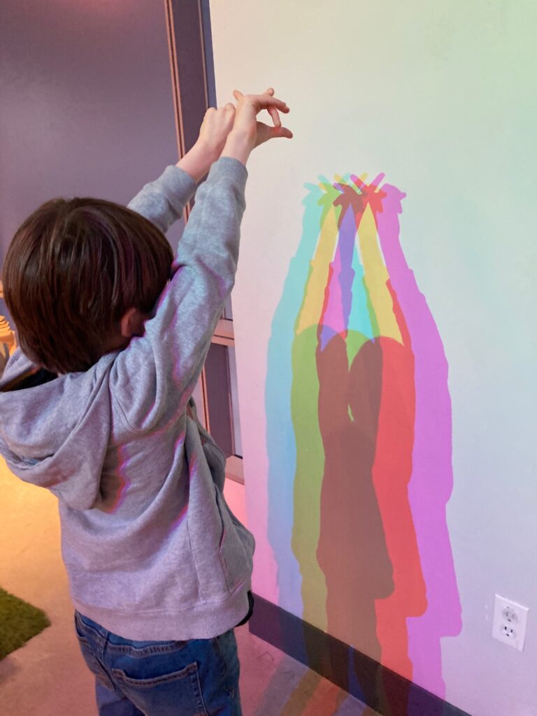 Photo of a young white boy holding up his hands above his hands, with a white wall showing a shadow split up in rainbow colors