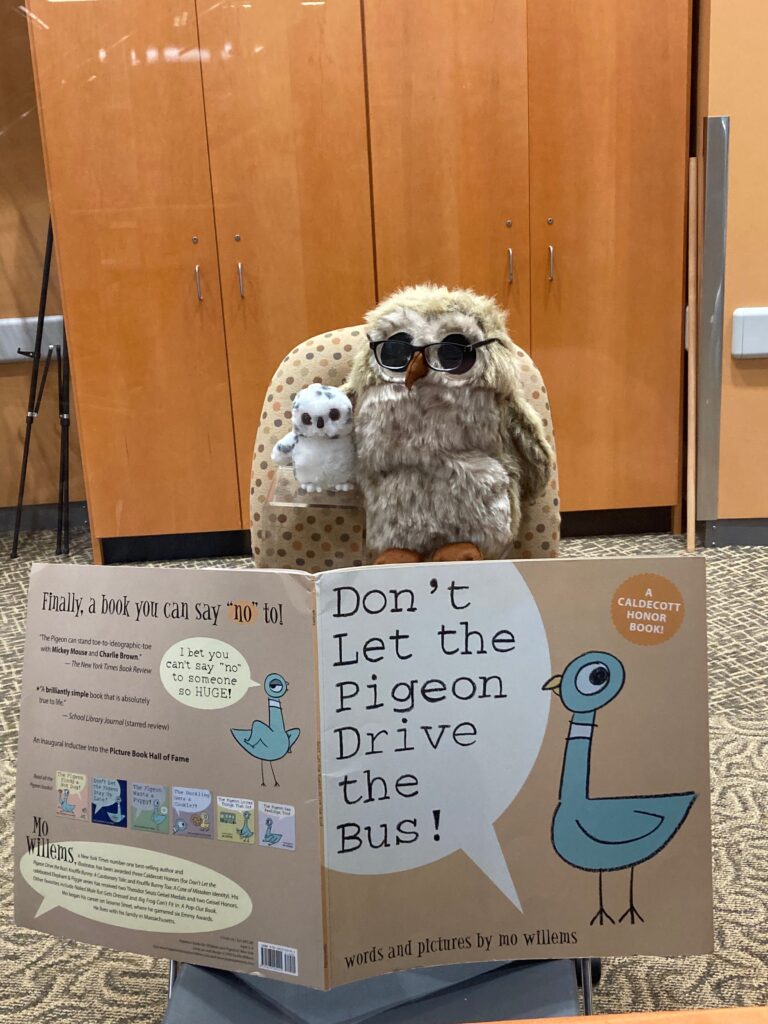 Photo of a large stuffed owl with glasses next to a small stuffed owl appearing to read a large version of a book Don't Let the Pigeon Drive the Bus!