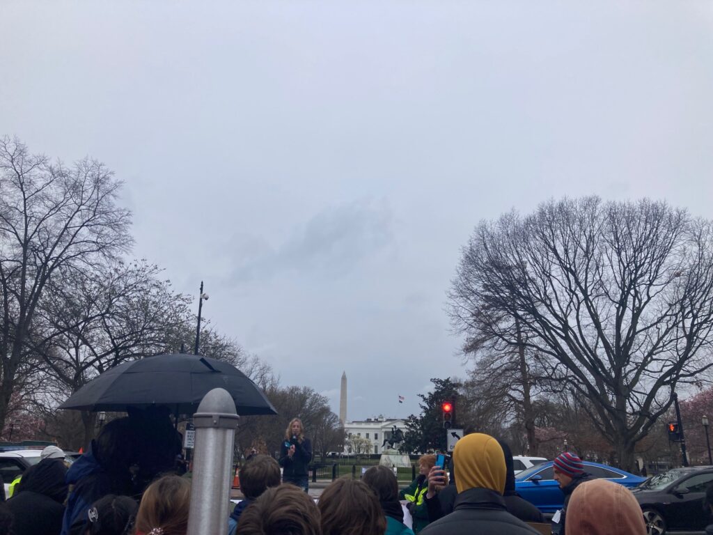 Photo of a climate justice protest in Washington D.C. with trees, the White House, and the Washington Monument behind a gathered crowd on a rainy day
