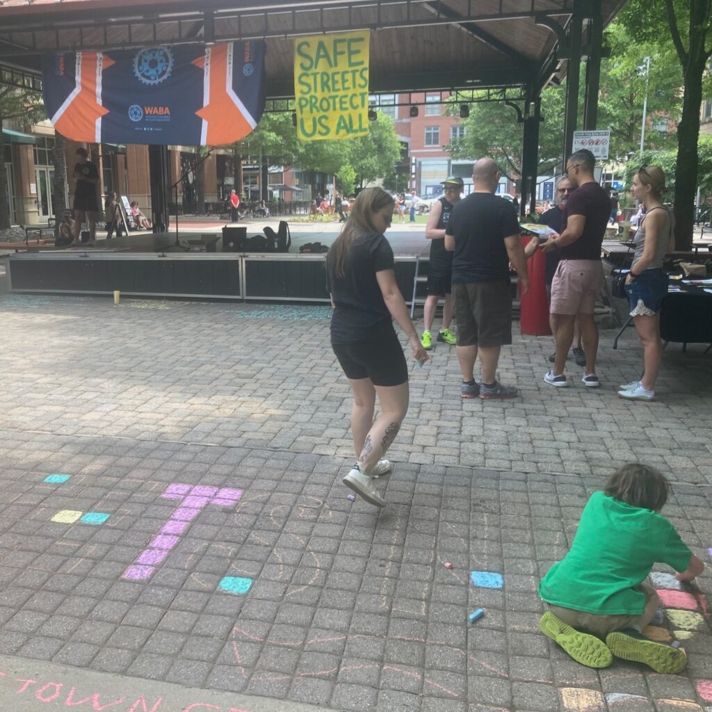 My older son (a white boy in a green shirt) leaning down and chalking, with another volunteer walking by and a group standing near a stage. There are two signs hanging from the stage, one of which has the WABA logo and one of which says "Safe Streets Protect Us All."