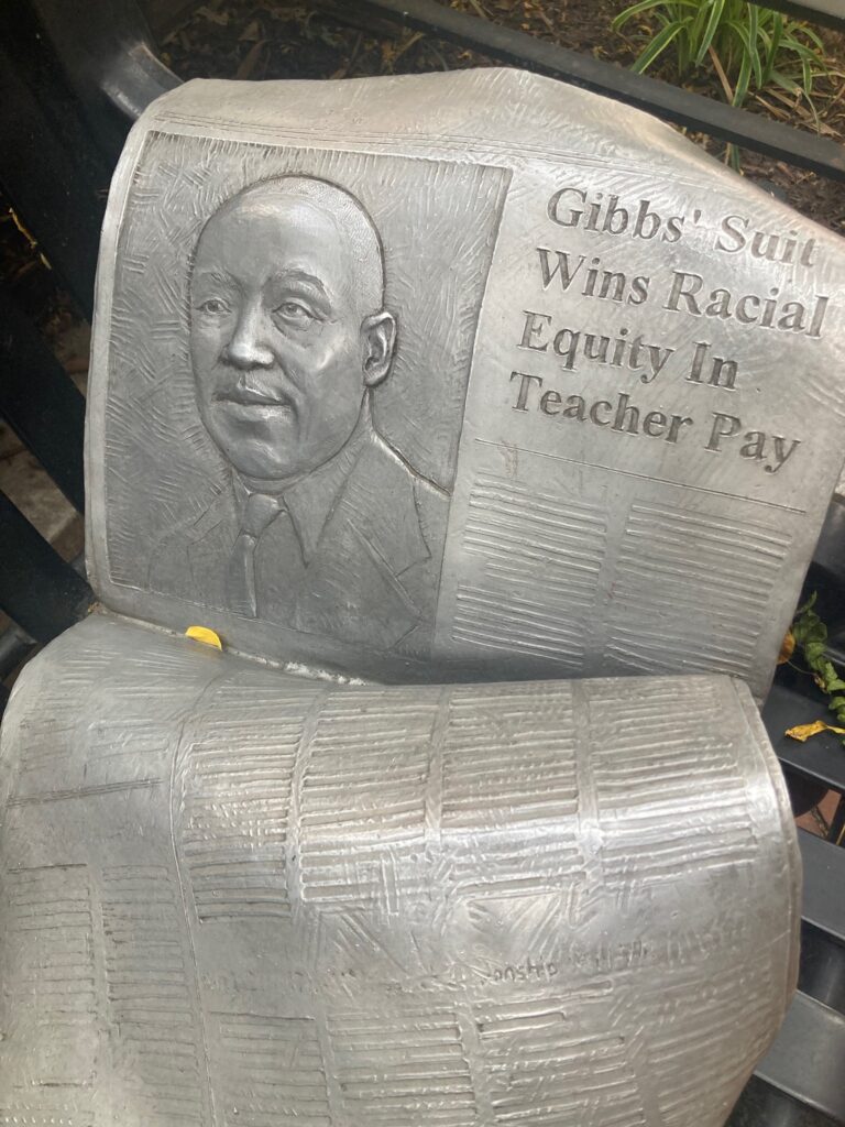 A metal statue of a newspaper with the headline "Gibbs' Suit Wins Racial Equity in Teacher Pay" with a picture of William Gibbs next to it 