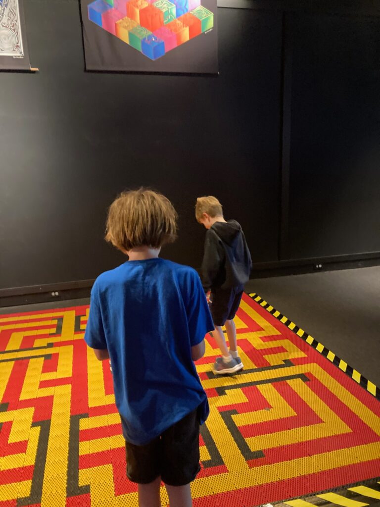 My kids (two white boys, one in a blue t-shirt and one in a black sweatshirt) walking on a maze printed on a giant carpet-like mat