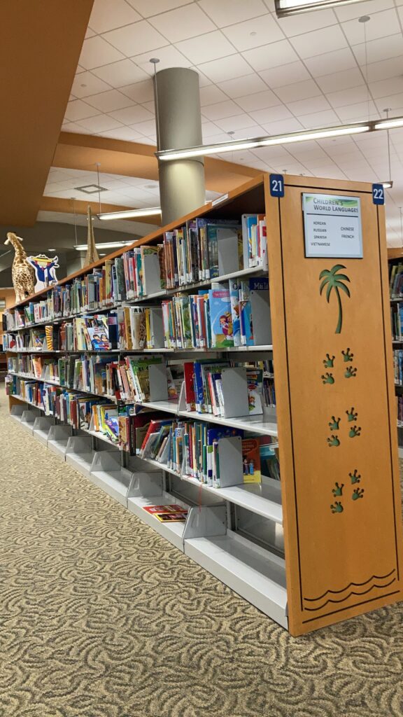 Photo of a bookshelf at our local library, filled with books, with a stuffed giraffe and a cardboard cutout of Madeline on top