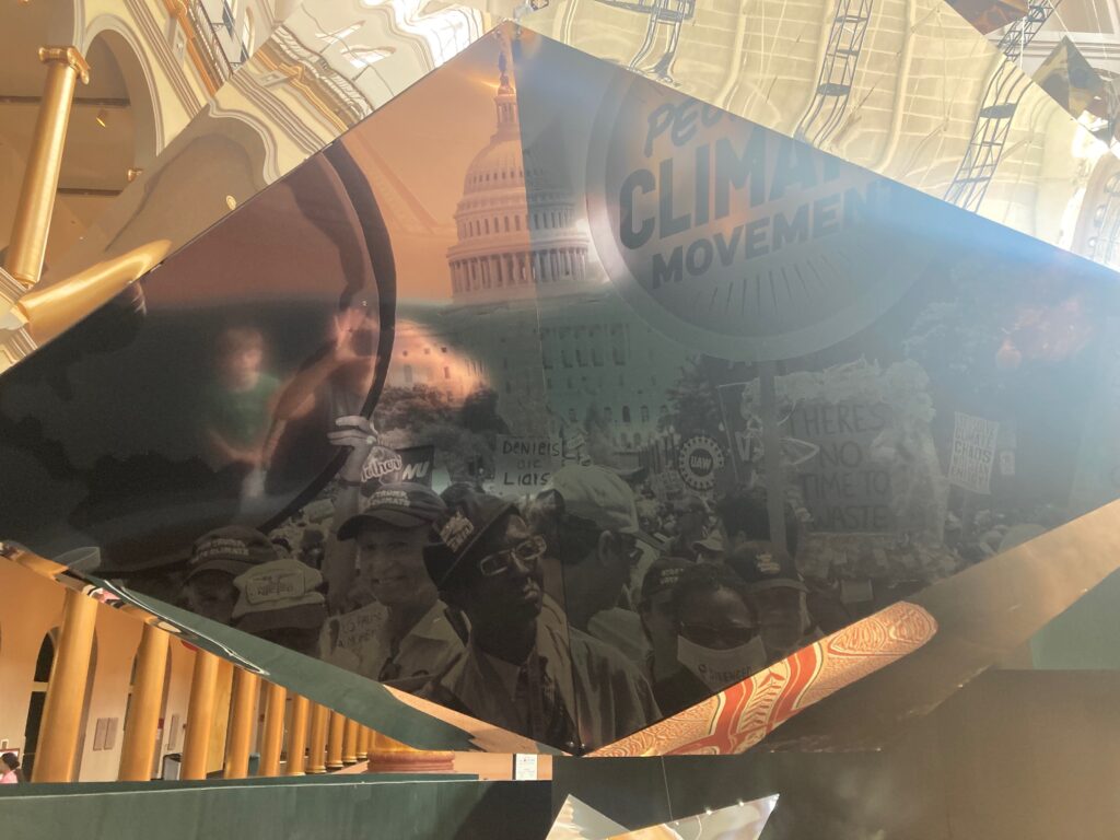 A shiny reflective object with a photo of the People's Climate Movement march with people of various races and ages holding signs in front of the U.S. Capital. A woman taking a photo and a child are reflected in the surface.