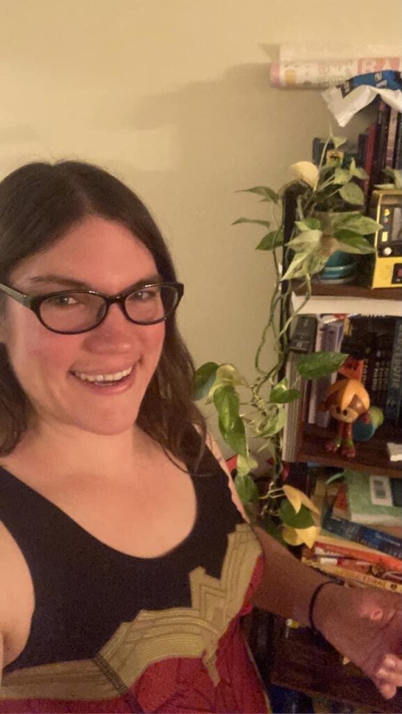 A photo of me (a white woman with brown hair and glasses) in a Wonder Woman dress standing in front of a bookshelf with books and a plant on it
