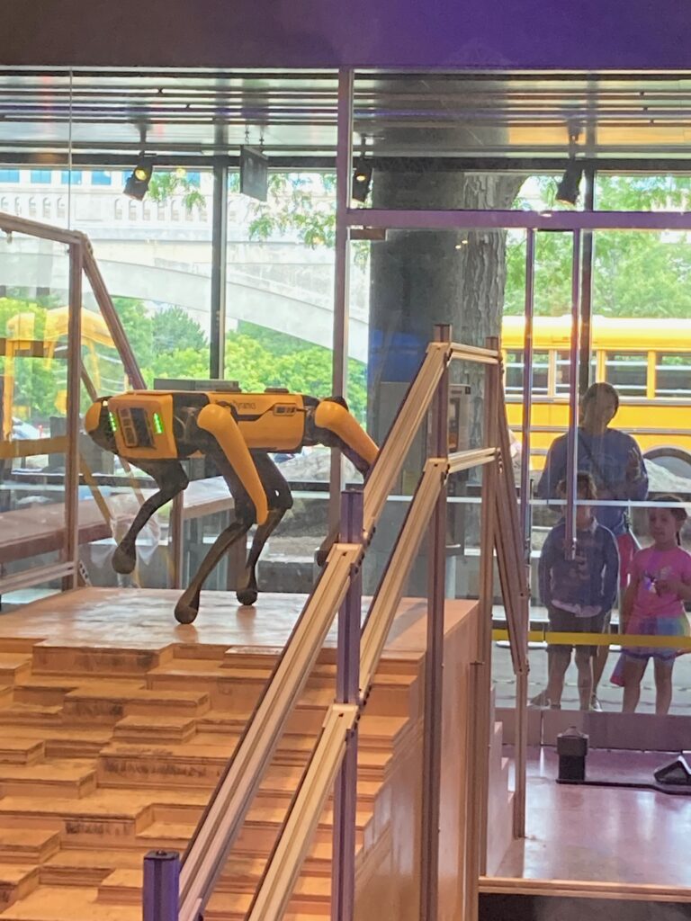 A robot dog that has a yellow and black body "standing" on top of a set of uneven stairs with two children and an adult looking through a window on the other side