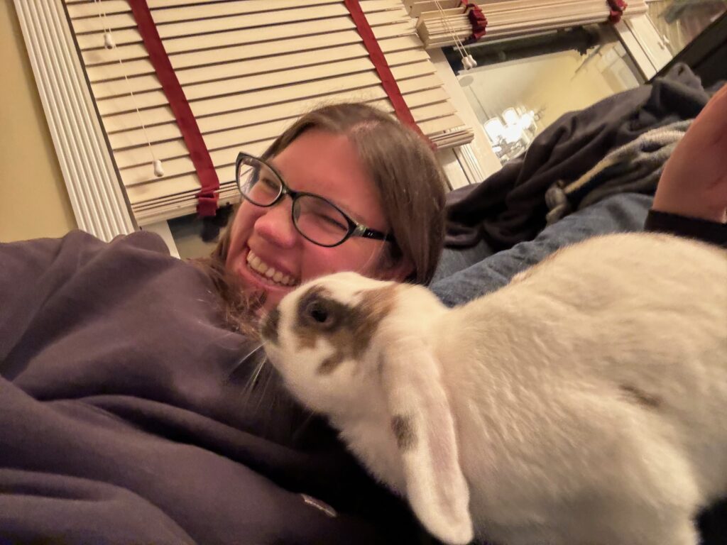 Me (a white woman with brown hair in a black hoodie sweatshirt) falling over laughing while having a white rabbit with brown splotches apparently jumping on me