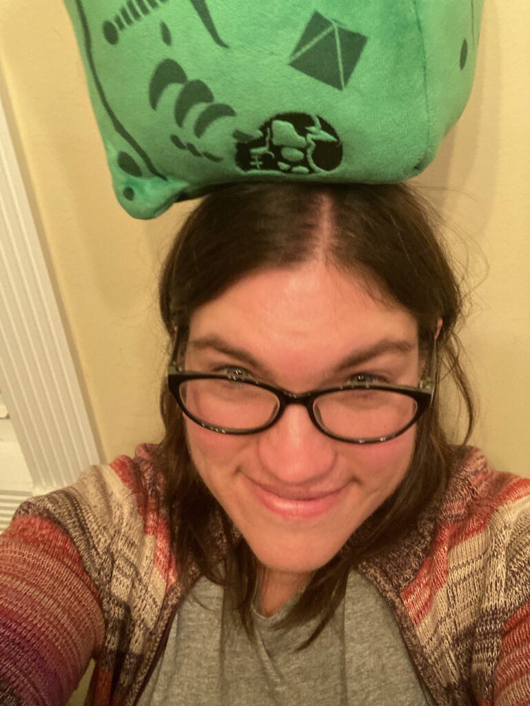 Me (a white woman with brown hair and glasses in a multicolored sweater) balancing a stuffed Gelatinous Cube on my head