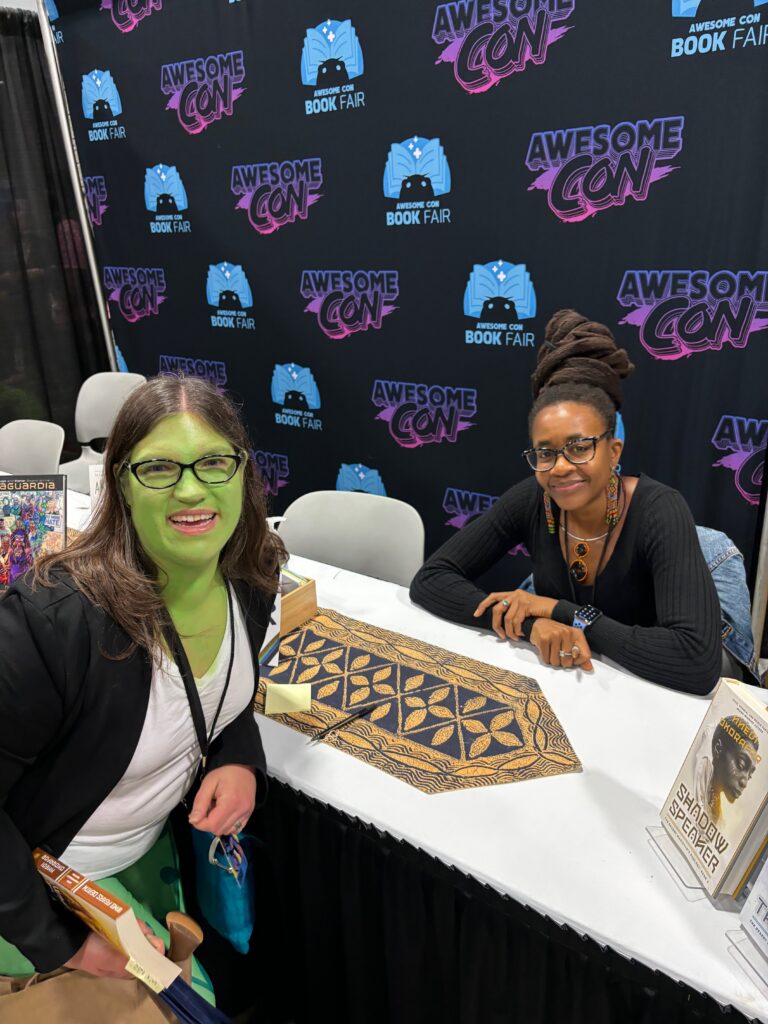 Me as Poison Ivy (a woman with green skin) from Harley Quinn posing with Nnedi Okorafor (a Black woman in a black sweater) at her signing with a large AwesomeCon backdrop 