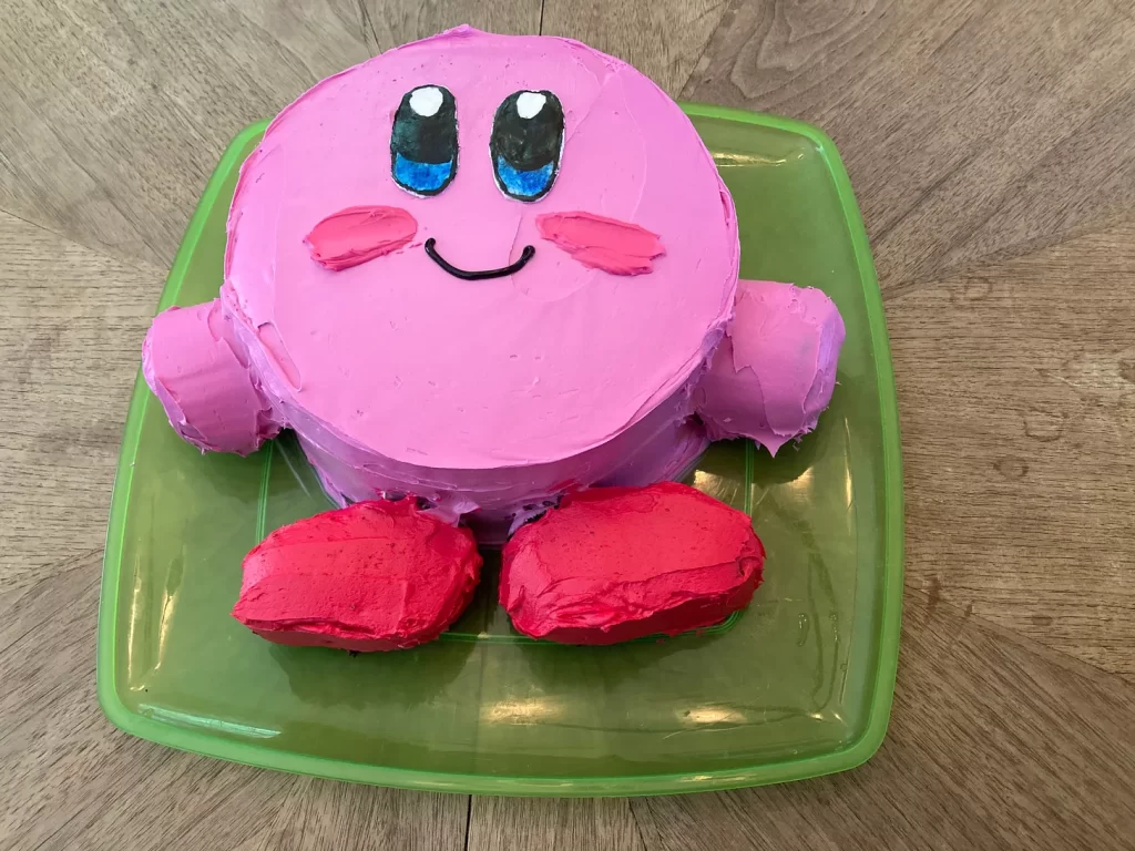 A cake shaped like the video game character Kirby, who is a round pink smiling ball with hands and feet.