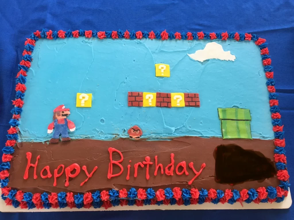 A sheet cake showing the first screen of Super Mario Bros, with Mario facing a Goomba, question blocks and bricks, and a pipe. It says “Happy Birthday” with the name edited out.