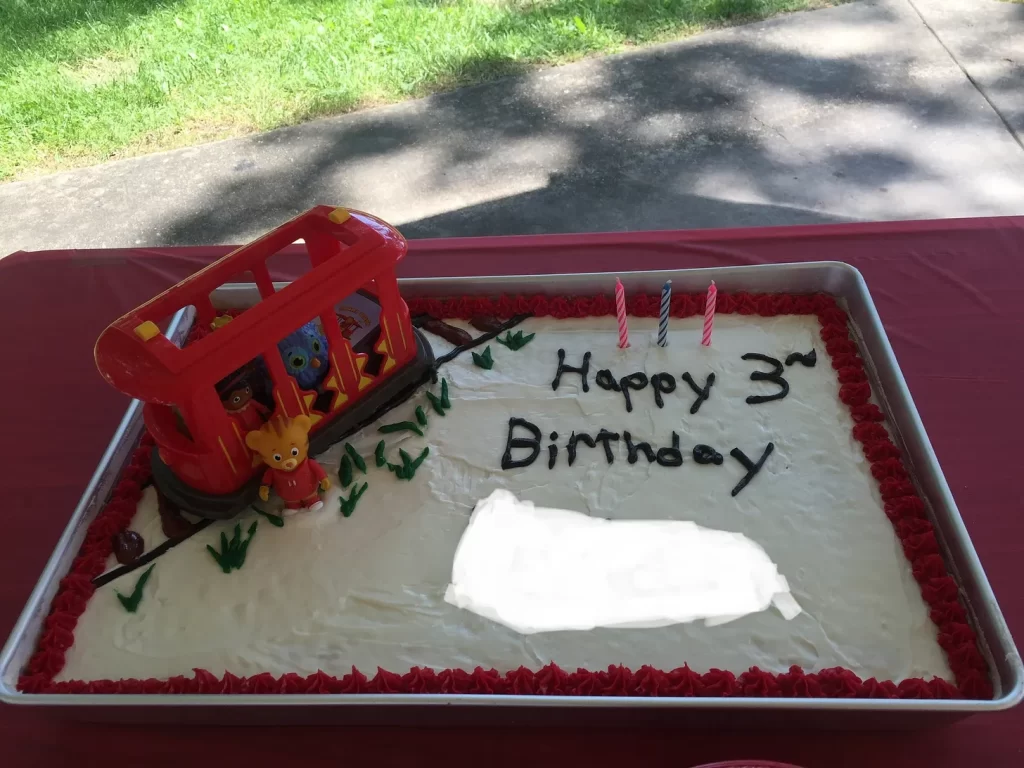 A sheet cake that says “Happy 3rd Birthday” with the name edited out. There is a Daniel Tiger figurine and a toy trolley on an icing trolley track with grass next to it