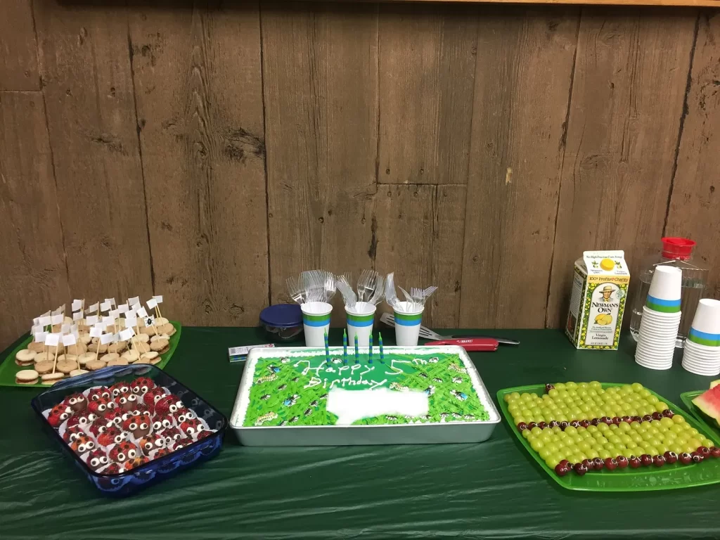 A table full of bug-themed food, with a sheet cake that says “Happy 5th Birthday” with the name edited out. It has lots of green grass with sugar bugs on it. There are also strawberry ladybugs and caterpillar grape skewers.