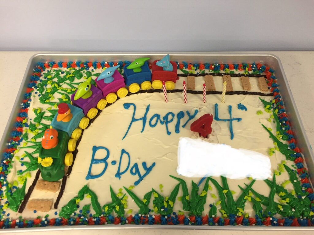 A sheet cake with the words “Happy 4th Birthday” and the name edited out. There is a train track curving around the edge with a Dinosaur Train toy on it. The tracks have frosting grass around them and a border surrounds the cake