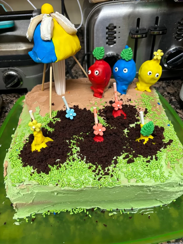 A square cake with grass frosting and a multi-colored orb and 3 little alien looking creatures lined up next to it. There are more “planted” inside the cake below them