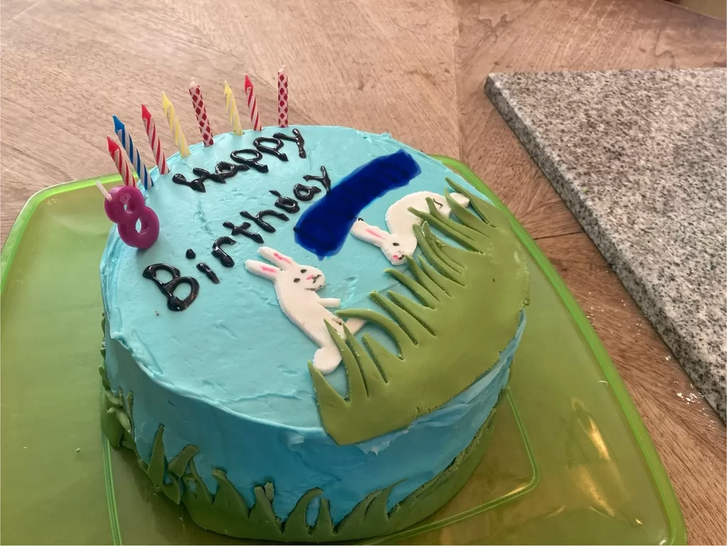 A round cake with green grass and a blue sky on both the top and sides of the cake. There are two white rabbits on the grass on the top, candles, and the words “Happy Birthday” with the name edited out