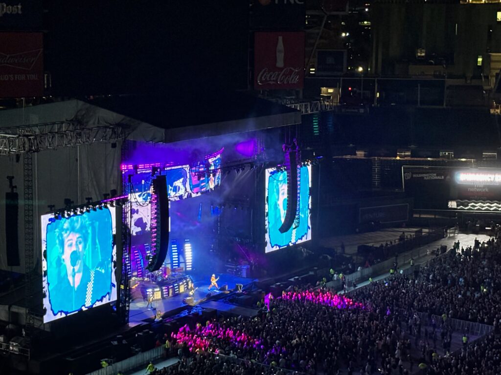 Concert at a baseball stadium, taken from high in the stands. Green Day are on stage, with singer Billy Joe's face projected on giant screens on either side. The area in front of the stage is packed.