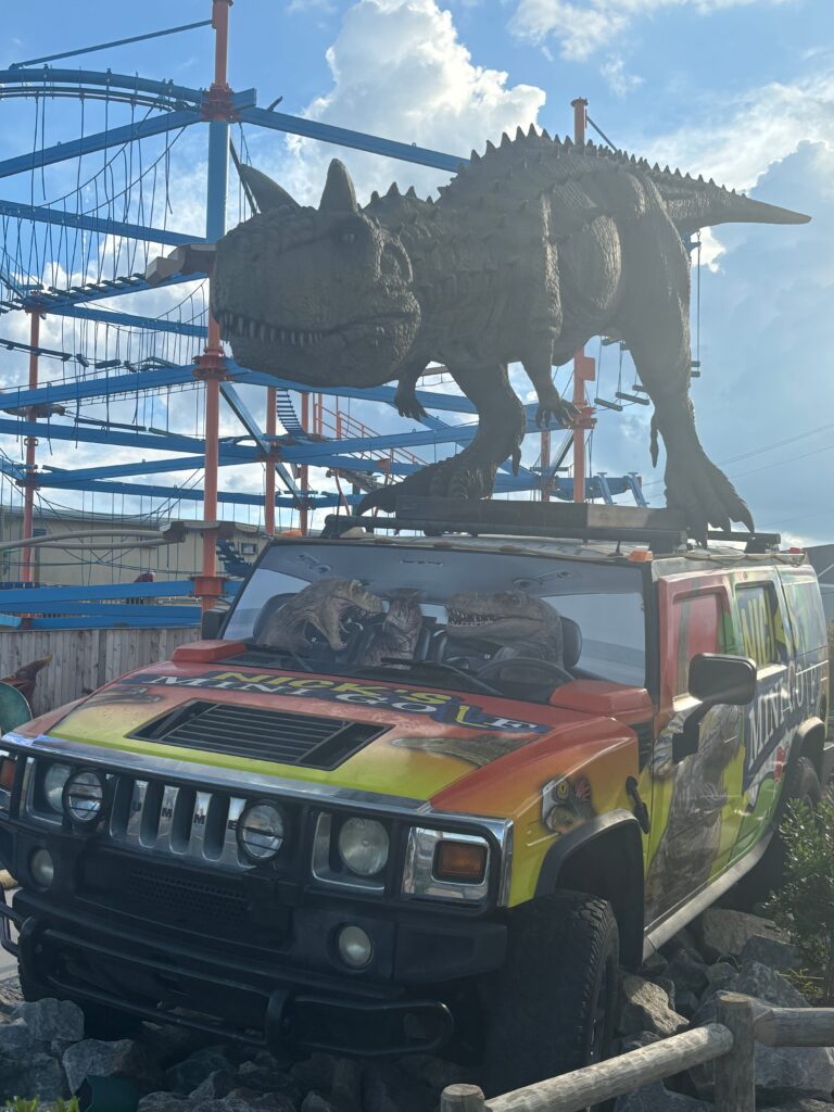 A statue of a T-rex on a Jeep (that has two velociraptors driving it) labeled with Nick's Mini-Golf. There is a structure for a high-ropes course in the background. 