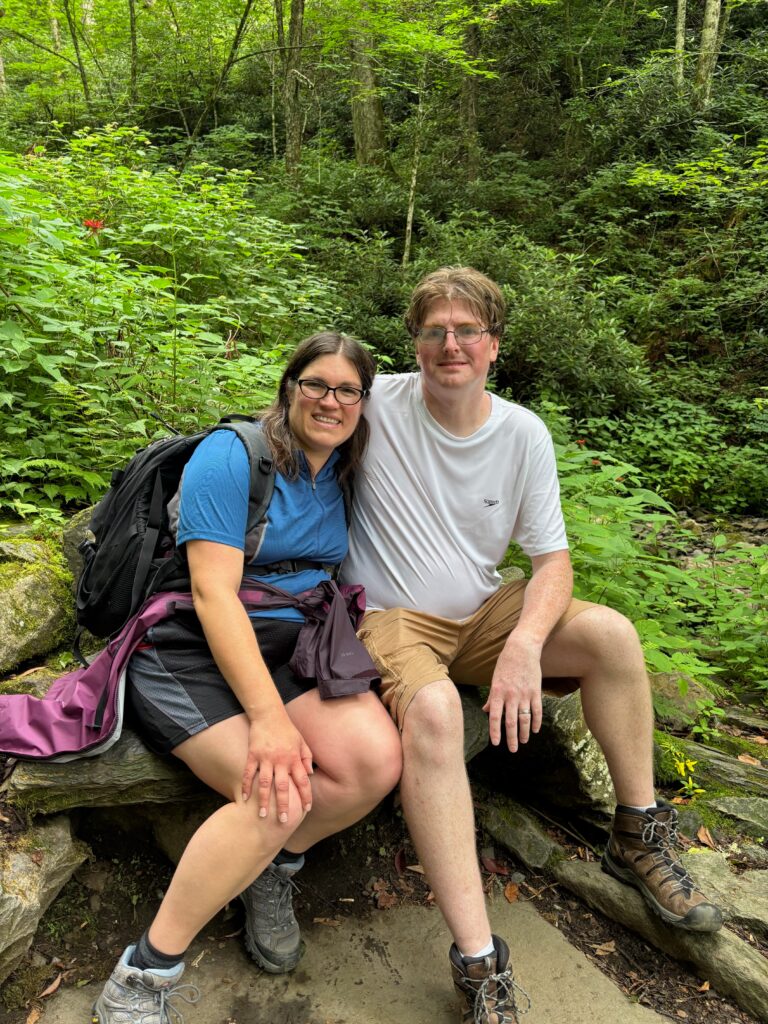 My husband and I, who are both white, in hiking clothing, sitting on a rock. I have a backpack on my back and there are trees and low plants in the background.