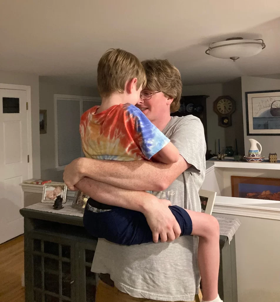 A white man (my husband) holding a white boy in a tie-dye t-shirt (my younger son) while standing in a living room